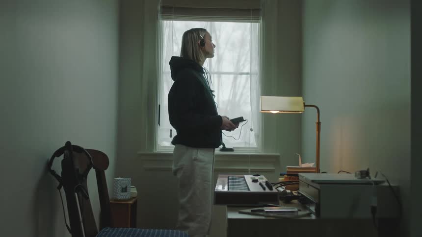 Blonde teenage girl in a hoodie standing beside a window with headphones on and a desk with a musical recording instrument in front of her