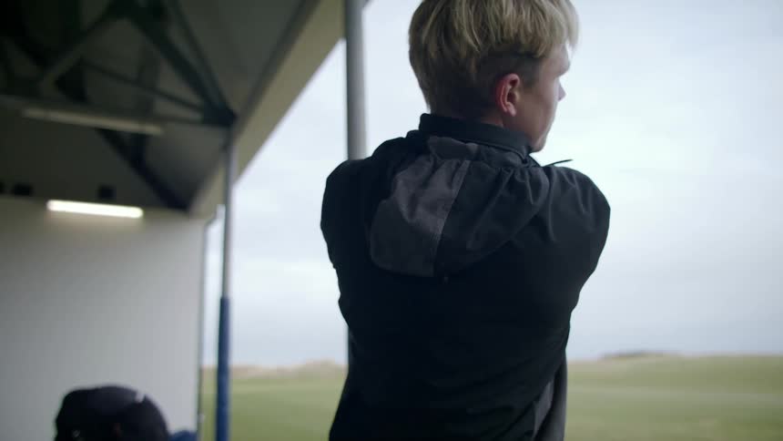 A blonde woman wearing a black jacket swings a golf club