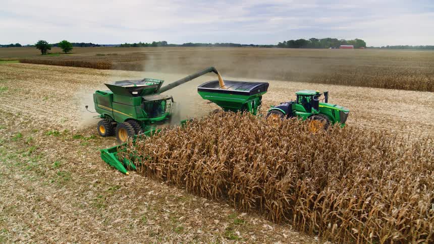Green john deere tractor pulling a harvest bin alongside a combine in a field collecting grain on a clear day