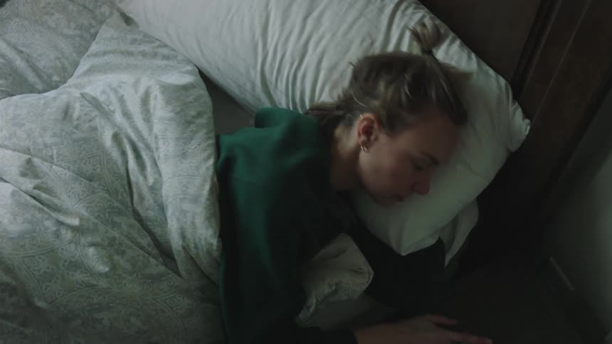 In the bedroom a woman in a green shirt and brown pants rises from the bed