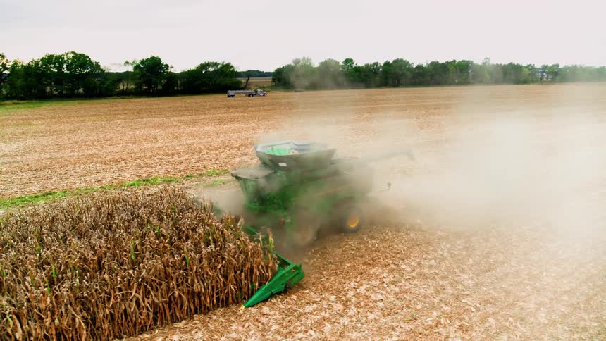 Drone profile shot of massive green combine harvesting rows of corn with dust kicking up