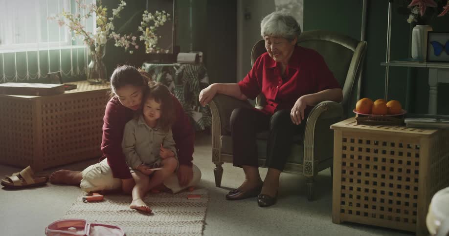 A sitting elderly woman watches as a young girl and her mother play on the floor in front of her