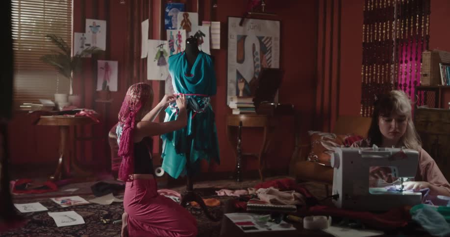 Two women sitting in a room one of them sewing and the other adjusting the clothing on a mannequin