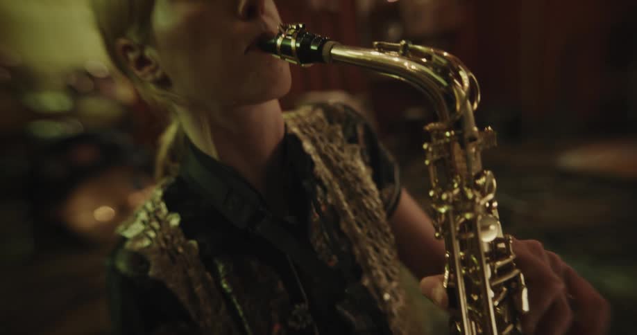 A young blonde woman sits in a room and plays the saxophone