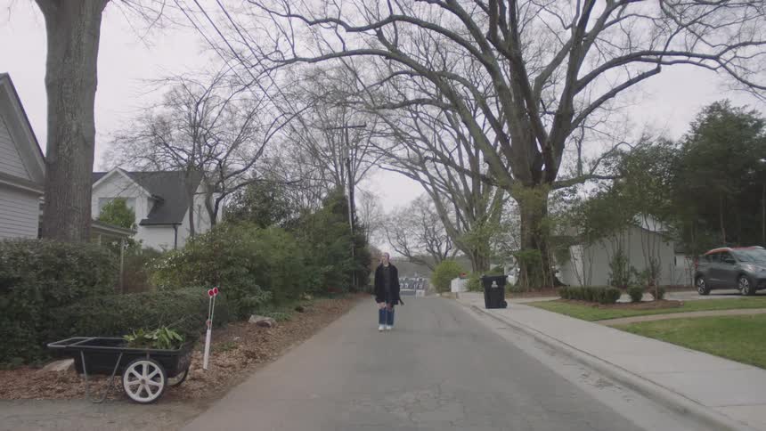 A teenage girl wearing torn jeans with a long coat and headphones walks on a neighborhood street