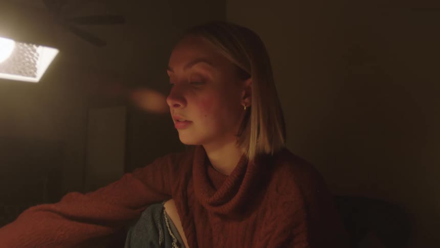 A tired woman with blonde hair and a brown sweater uses a smartphone next to a lamp in a room
