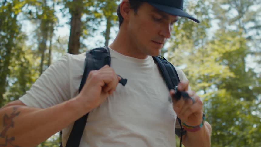 A man standing in the woods puts on a backpack and walks away