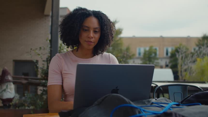A young woman sits outside on a patio and works on a laptop