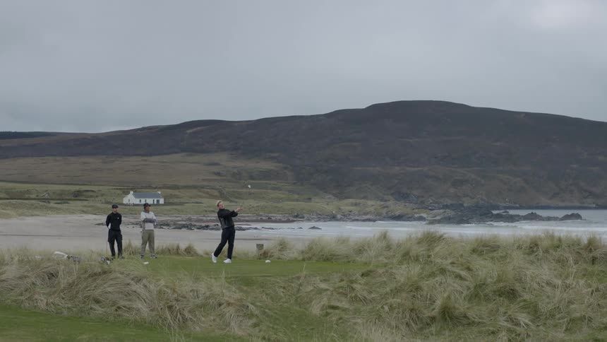 Three men on a seaside golf course with one of them taking a swing on a cloudy day