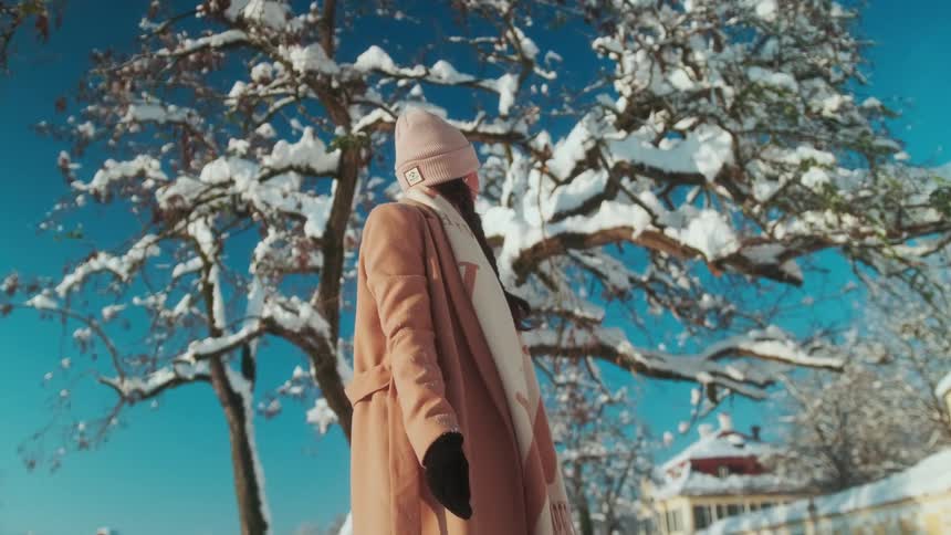 A teenage girl standing on a neighborhood near a snow covered tree as she turns and looks toward a building
