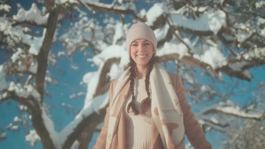 A young woman stands near a snow covered tree and smiles as she throws snow from a branch