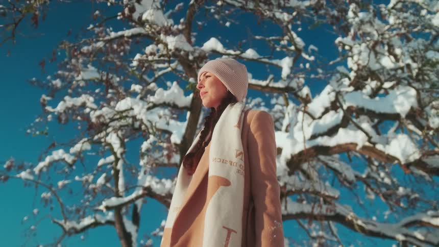 Woman in a winter coat and accessories standing outside in the sun on a winter day with snow covered tree branches and a blue sky in the background