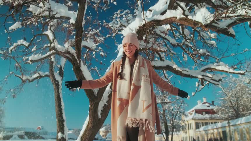 A young woman standing under a snow covered tree reaches up to the branches and smiles as snow falls down as she walks away