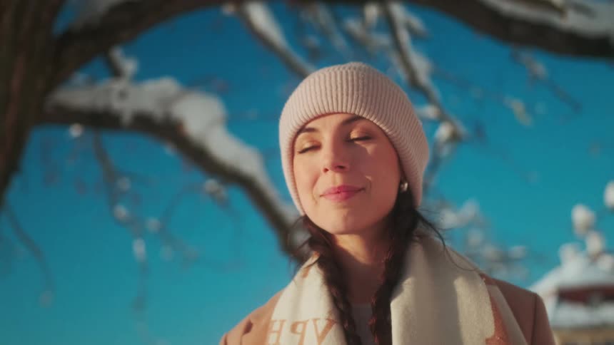 A happy woman with a white beanie braids and a brown coat stands under a snow covered tree embracing the wintry scene