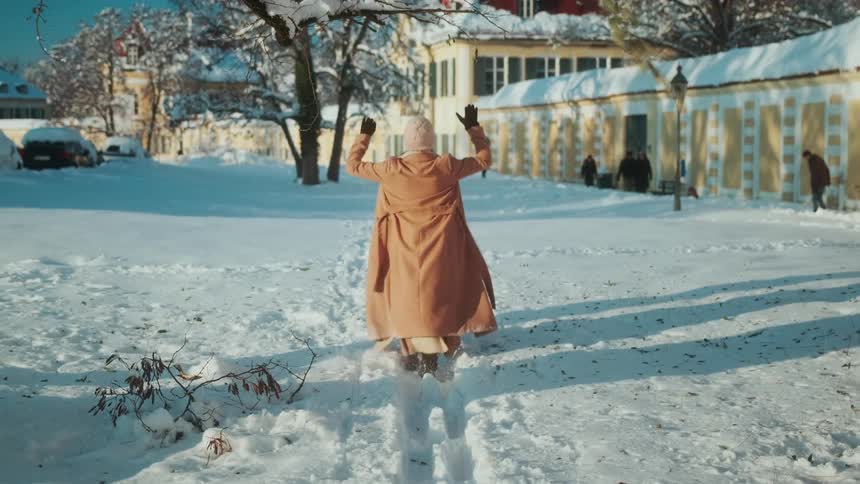 Woman runs through snow then breaks and throws tree limb as people walk near building in the background