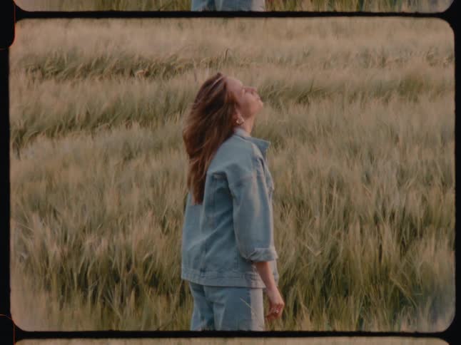 A woman with red hair wearing a denim jacket and jeans stands in a wheat field