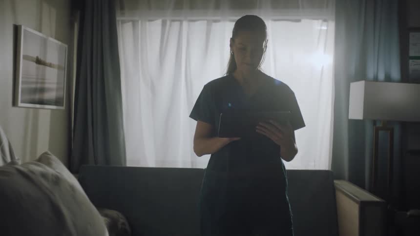 Nurse wearing scrubs enters data on tablet while standing near hospital bed