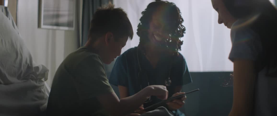 Nurses smile while they sit and let young male patient play on their tablet in hospital room