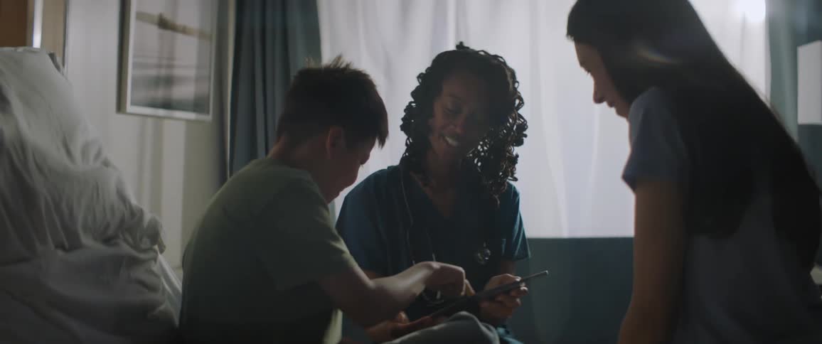 Clinician and boy gesture over tablet female sits to the side indoors