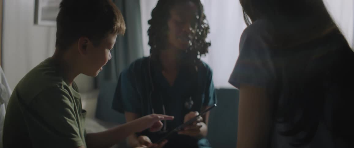 Woman in scrubs talks to boy gesturing on tablet sitting in clinic