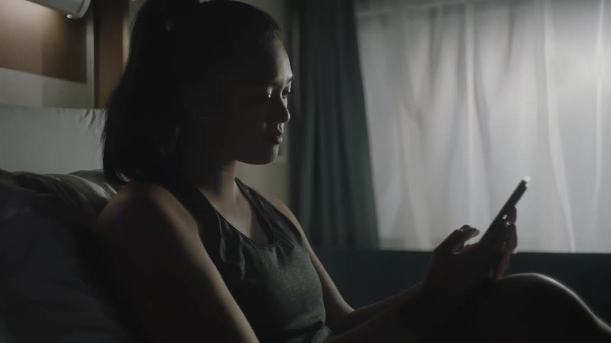 Asian female sits looking at cell phone in front of window indoors