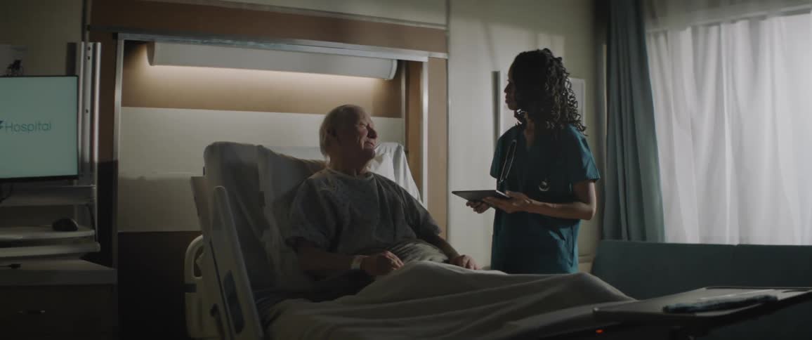 Woman in scrubs stands near elderly patient in hospital bed