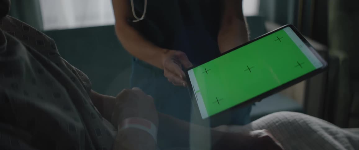 Nurse holding a tablet device in front of a patient in a hospital bed who is pointing to the large plus signs on the green screen