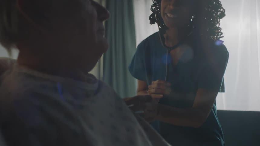Two young friendly healthcare workers smile and chat with an elderly male patient as they check his heart with a stethoscope