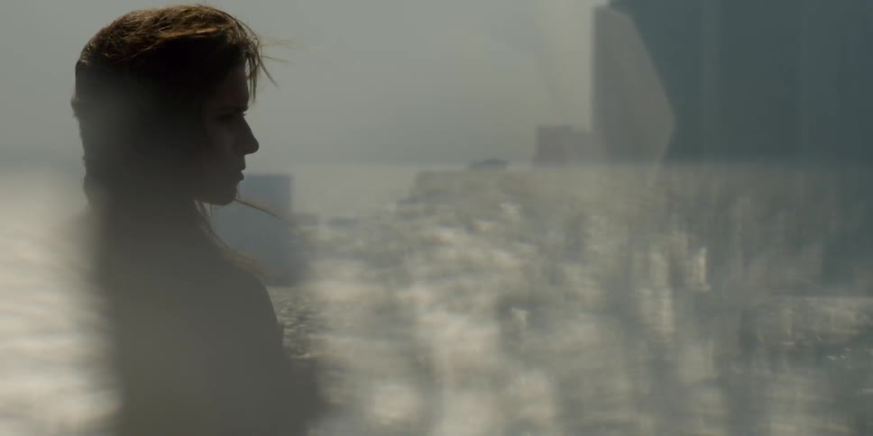 A woman stands near the shore as the wind blows her hair and the sunshine glistens on top of the water