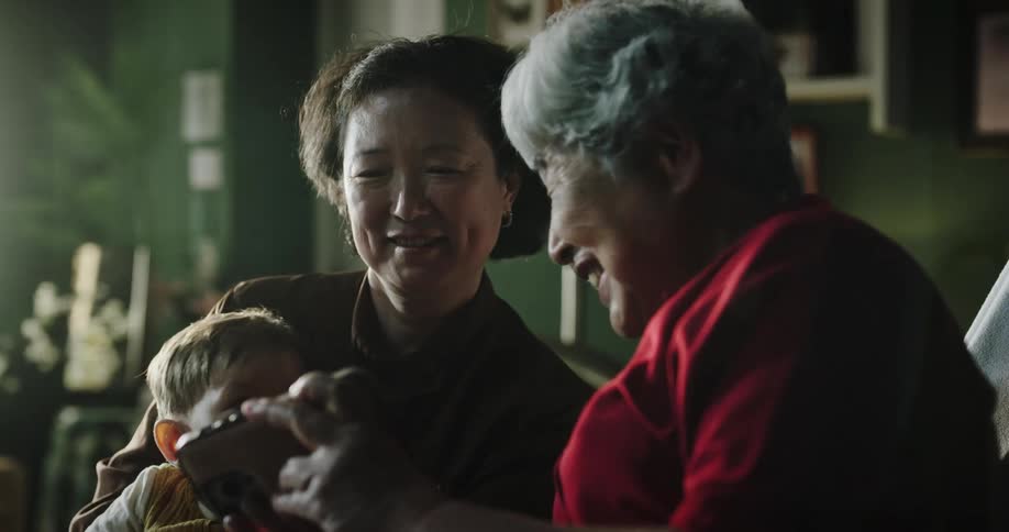 A family of a lady and a woman with a little boy on their lap and are looking at a cell phone and smiling