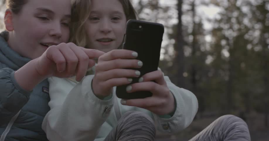 Two young children play with their phone outdoors near many trees