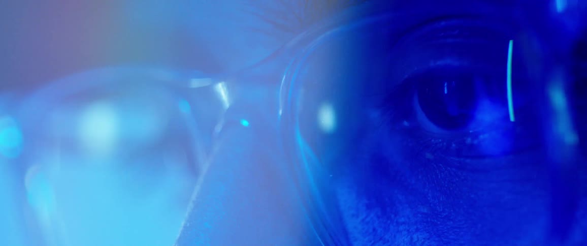 A person wearing glasses stares ahead while illuminated with blue light
