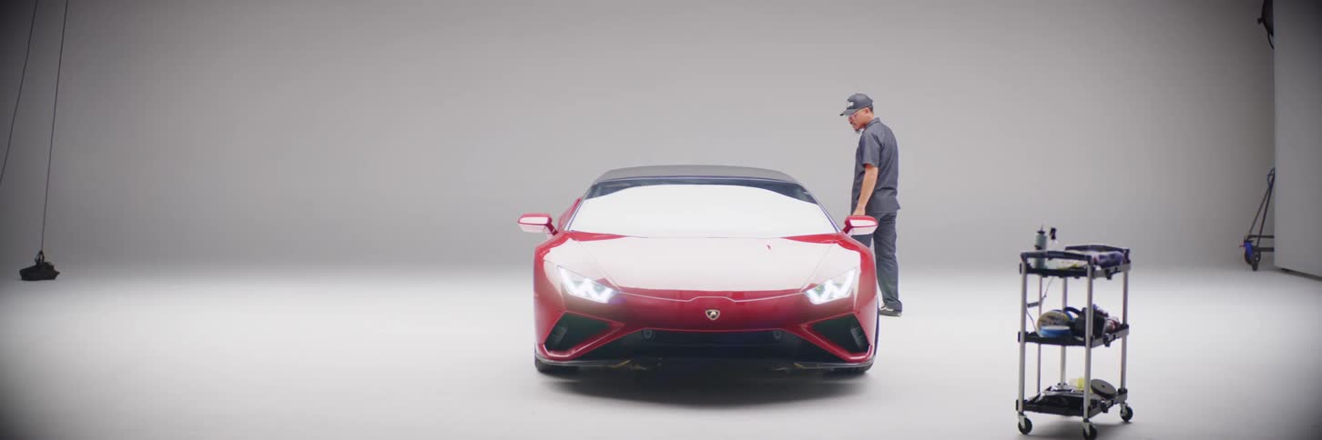 A man is walking and looking around a red sports car that is parked in a photo studio with a white background