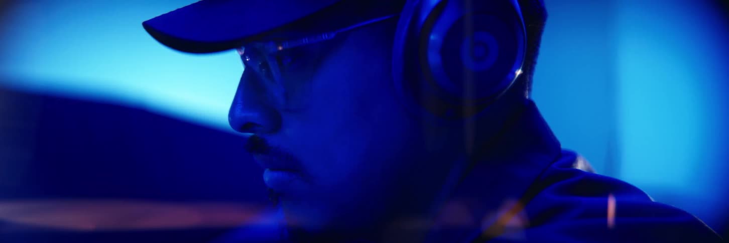 A man with glasses and headphones wearing a hat and looking straight ahead and the whole environment has blue lights