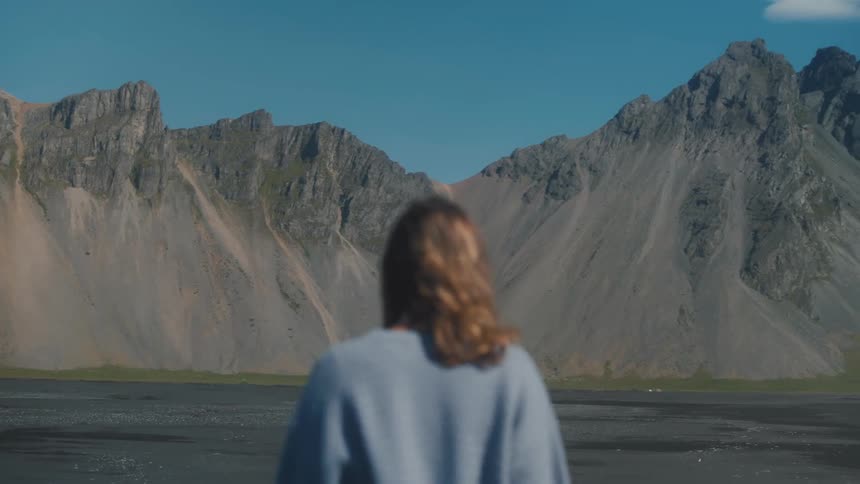 Woman walking towards a valley with black sand on a day with a blue sky