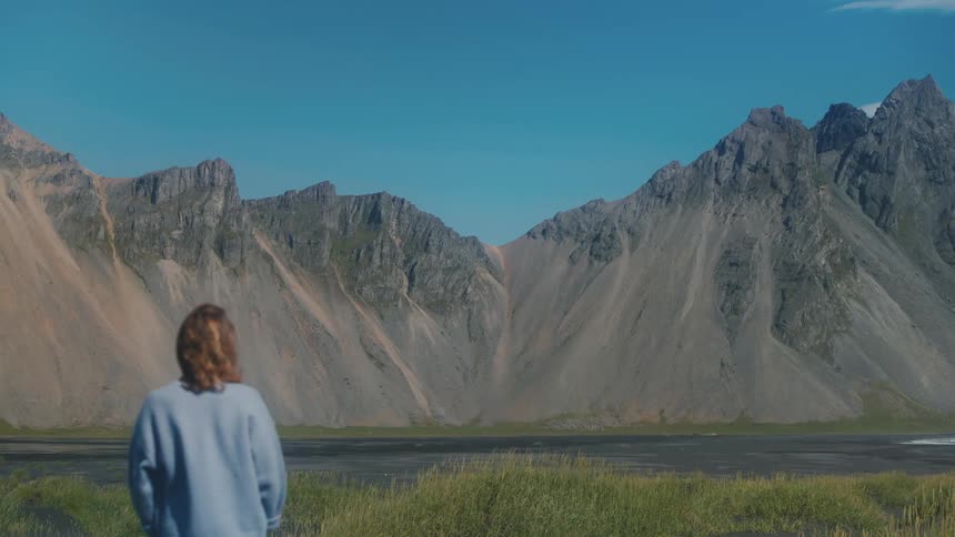Woman with her back to the camera and facing a lake with mountains around taking a photo with her phone on a day with a cloudless blue sky