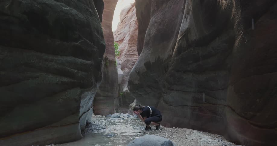 Woman in the midst of a canyon drinking water from a small flowing river while it drizzles