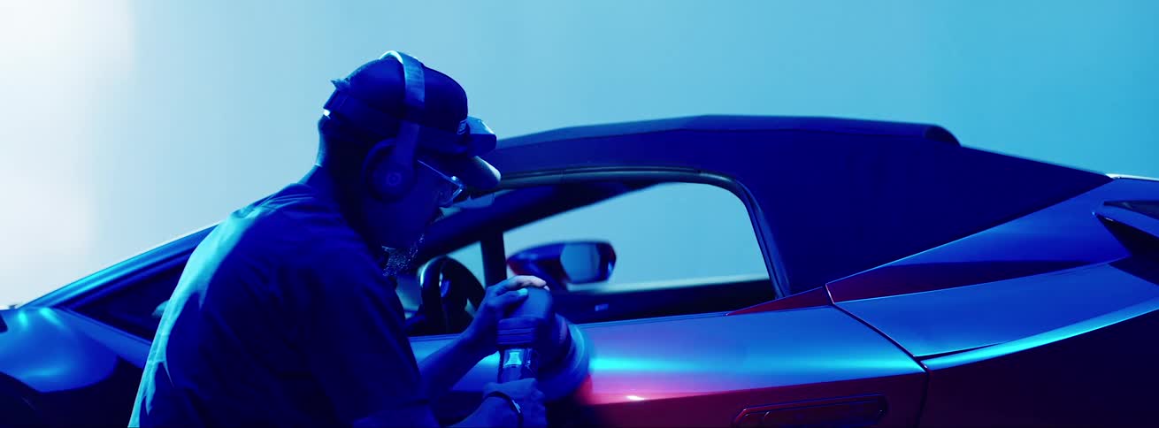 A man buffs an expensive car while spotlights dance behind him against a blue background