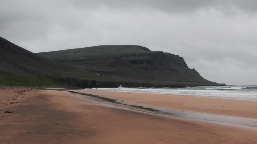 Waves roll into shore and over the beach near a promontory under a cloudy sky