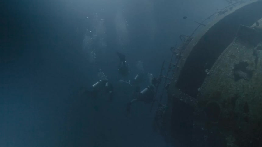Scuba divers swimming in cloudy blue water near a sunken ship resting on its side at the bottom of the ocean