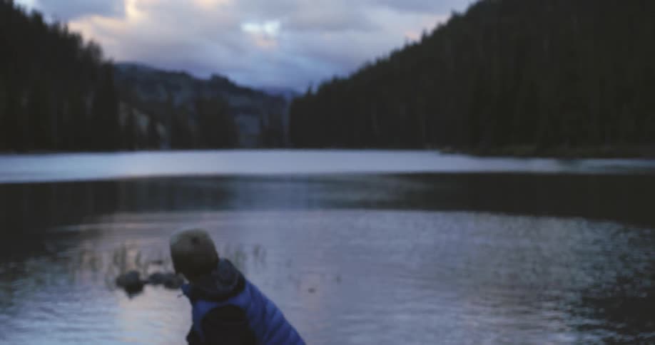 A boy throws a rock into a lake surrounded by hills