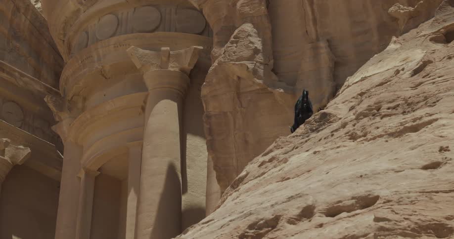 A black goat is on top of a mountain of stones that is next to an architecture carved into the stones