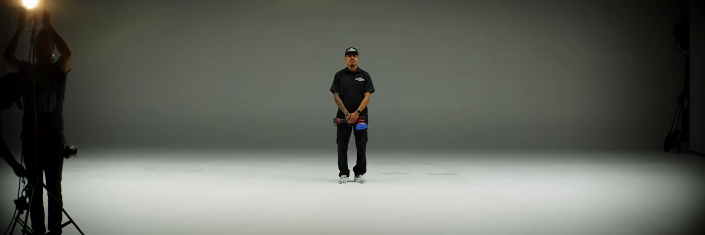A man wearing a cap and a cloth in his hand is standing in a photo studio looking forward and the background behind him is all white