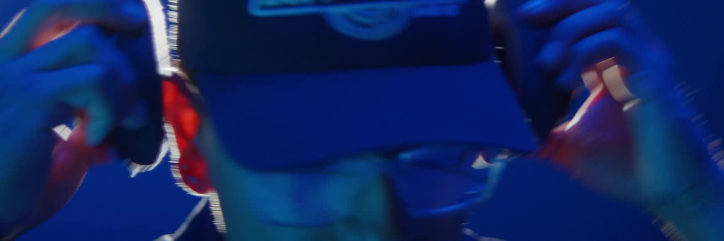 A man with glasses and a hat puts a headphone on his head and the environment around him is blue