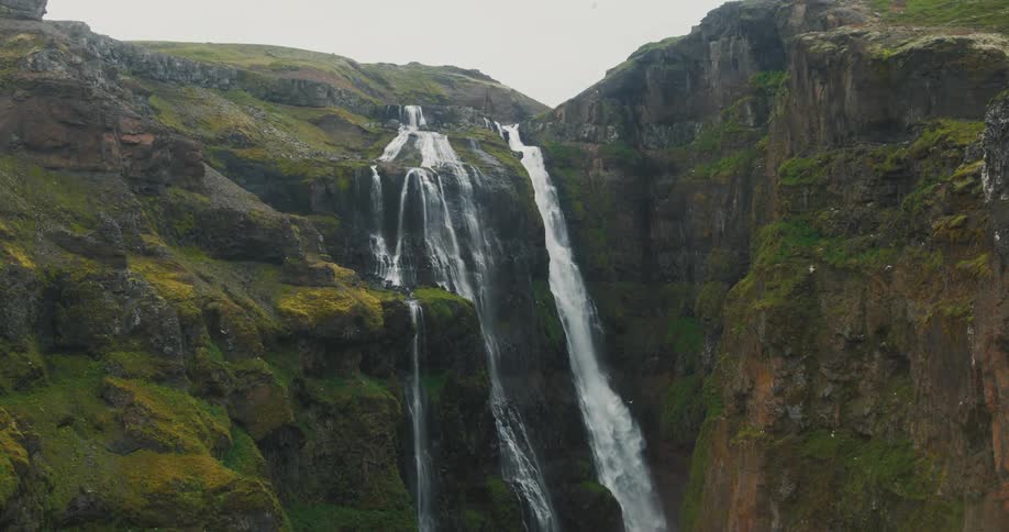 Waterfall dropping from a moss covered rock cliff into a valley carved out below on a cloudy day