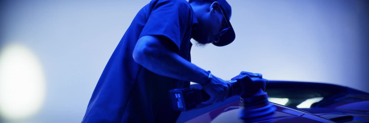 A man is polishing his car while spotlights dance behind him and everything is tinted blue