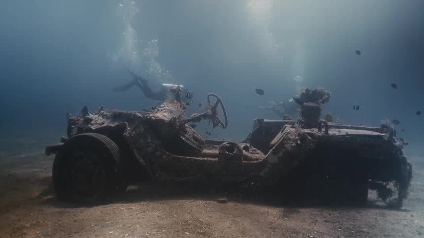 Two divers at the bottom of the ocean with a submerged car and fish around