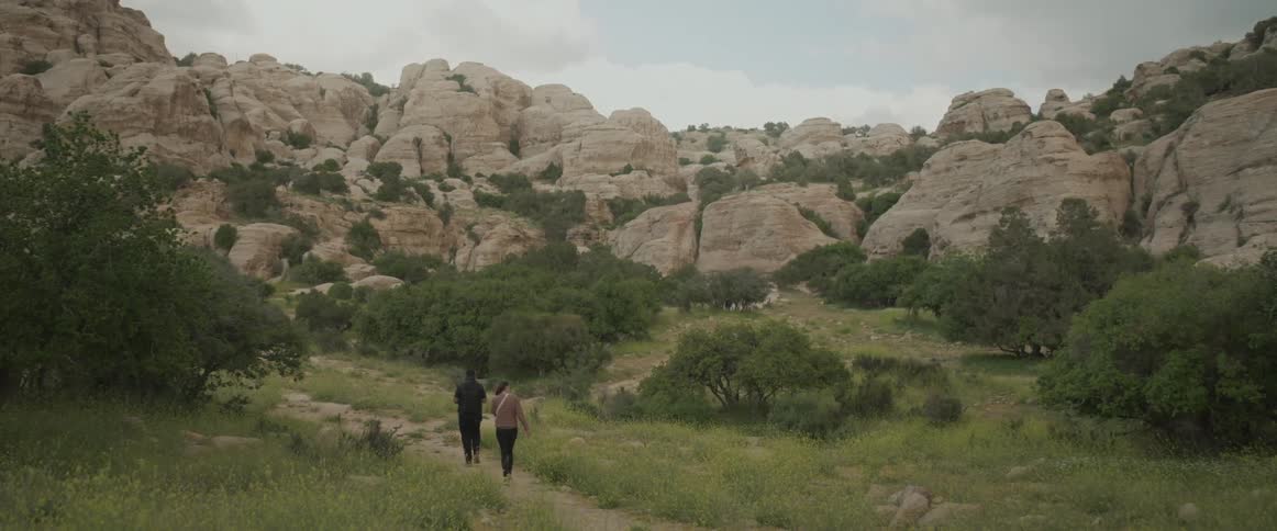 Two people walking on a path across a grassy field towards several large rock formations