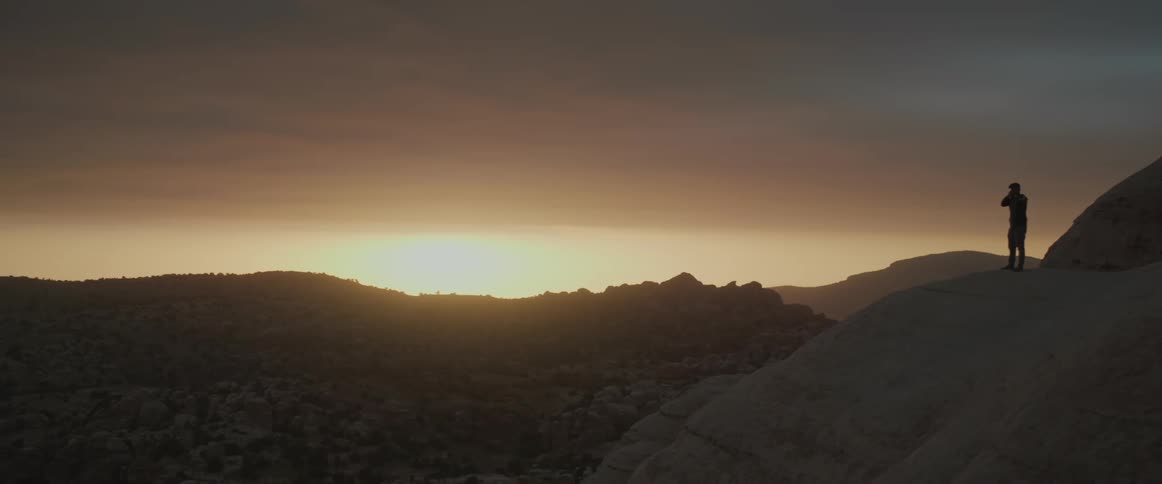 A man walking amidst cliffs under the mountains with the sunrise on the horizon