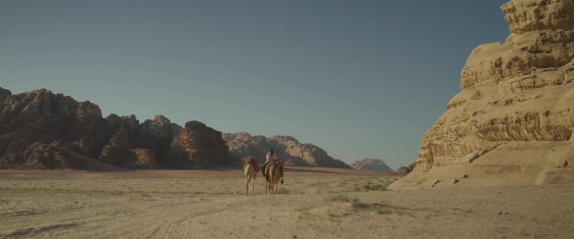 A person riding a camel walking through the desert surrounded by mountains